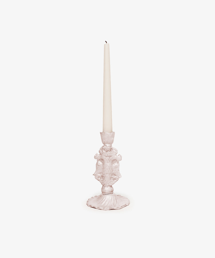 A white taper candle rests in the Aguila Bicefala Candleholder, an ornate, hand-moulded light pink piece, set against a plain white background.