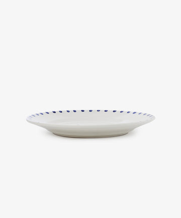 The Nina Serving Plate is a white ceramic plate featuring a blue dotted rim, shown from the side against a plain white background.
