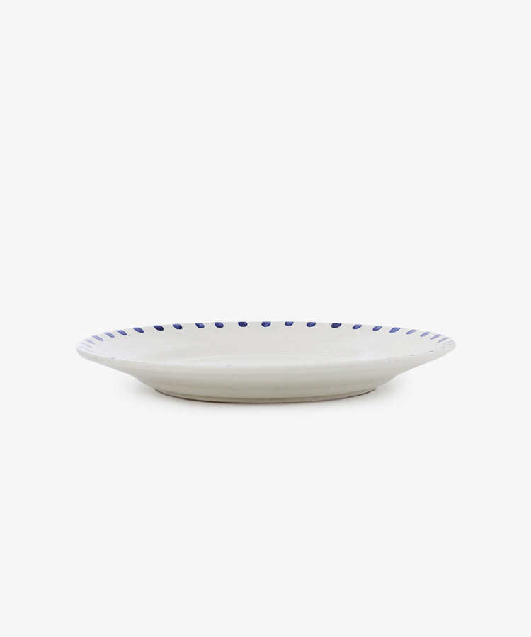 The Nina Serving Plate is a white ceramic plate featuring a blue dotted rim, shown from the side against a plain white background.