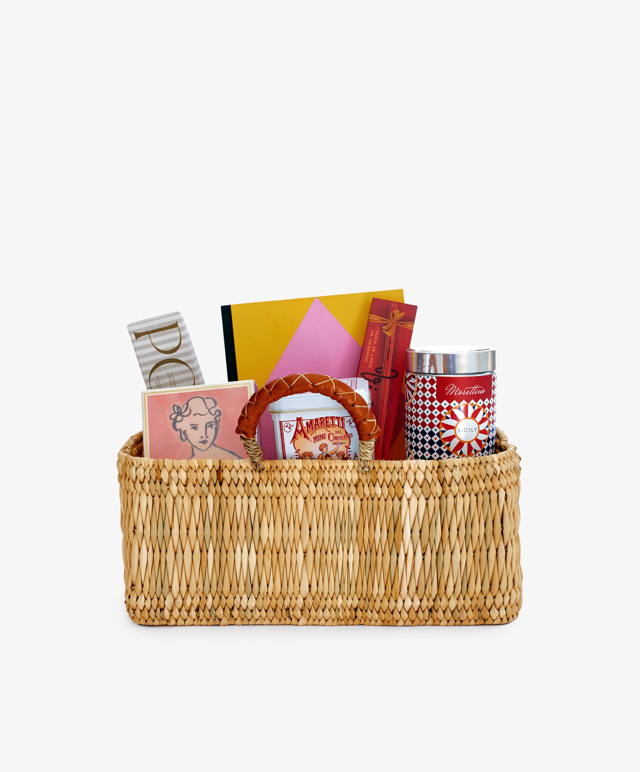 The Notebook & Nibbles Set is a rectangular woven desk gift basket filled with books, stationery, a tin, and assorted boxes, all neatly arranged against a plain white background.