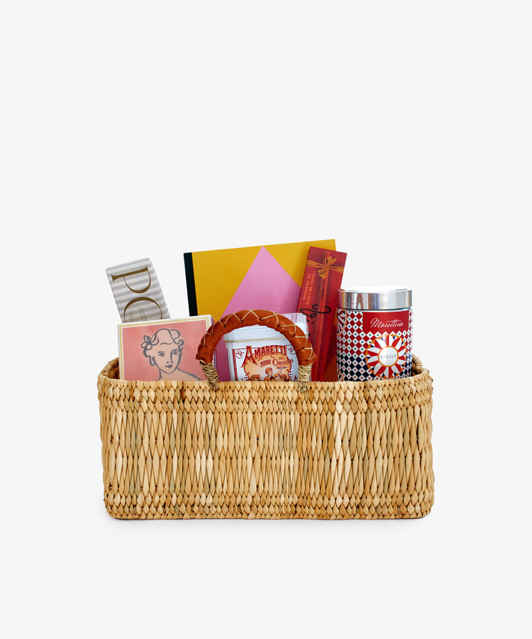 The Notebook & Nibbles Set is a rectangular woven desk gift basket filled with books, stationery, a tin, and assorted boxes, all neatly arranged against a plain white background.