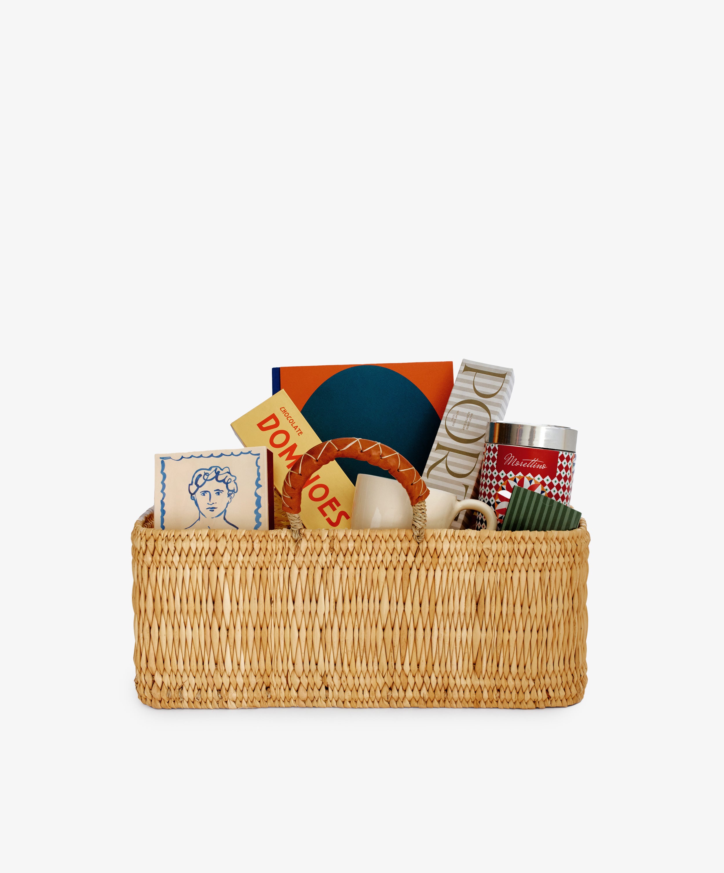The Gentleman's Edit: a woven hamper featuring a mug, luxury coffee, candle, chocolate domino set, tin, books, and a blue & white decorative box—all elegantly arranged upright against a white background.