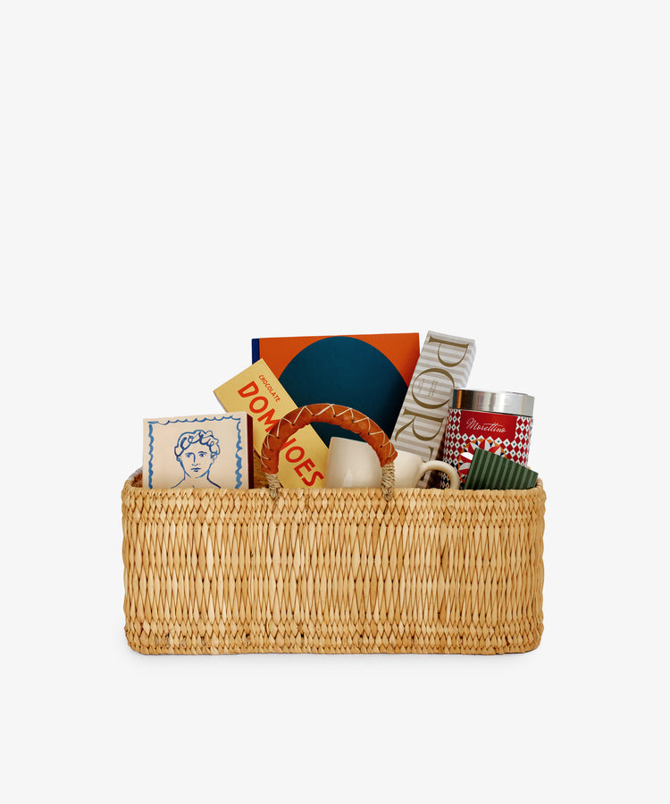 The Gentleman's Edit: a woven hamper featuring a mug, luxury coffee, candle, chocolate domino set, tin, books, and a blue & white decorative box—all elegantly arranged upright against a white background.