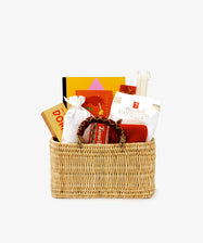 The Valentine's Hamper features books, packaged food, a white sack, two white candles, and a jar arranged against a plain background—an ideal gift for Valentine’s Day.
