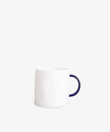 The Tea Mug features a minimalist plain white ceramic body with a dark blue handle, handcrafted quality, and is shown against a light gray background.