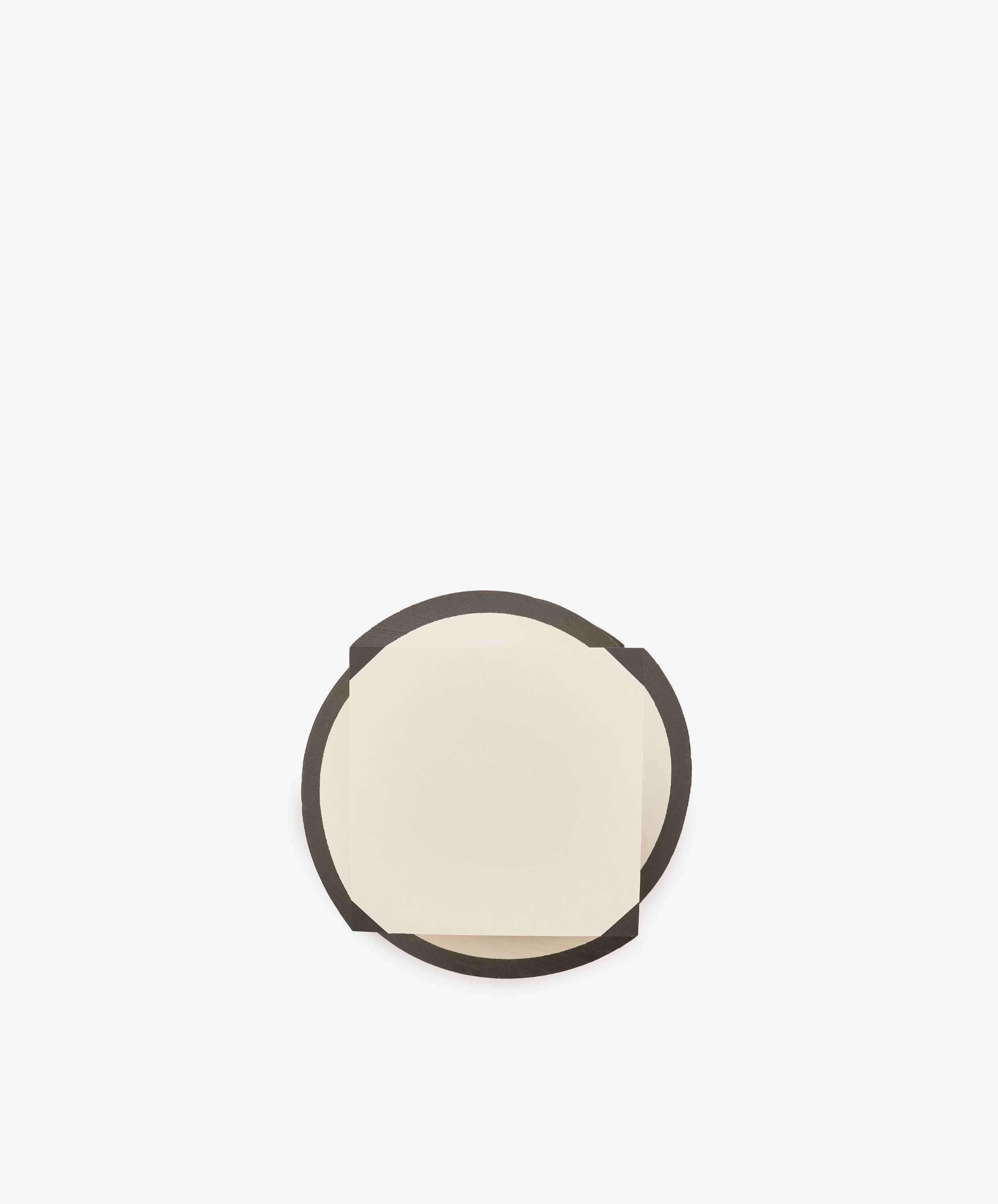 A small, square, cream-colored sheet from the Spiral Notepad sits on top of a larger, round, dark brown coaster against a plain white background.