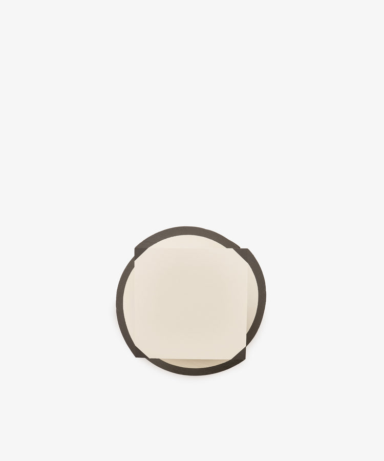 A small, square, cream-colored sheet from the Spiral Notepad sits on top of a larger, round, dark brown coaster against a plain white background.