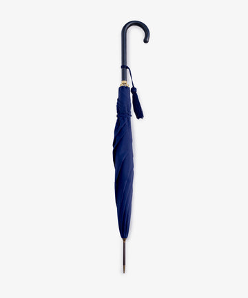 The Victoria Ladies Umbrella, a closed navy English umbrella with a leather crook handle and decorative tassel, stands upright against a plain white background.