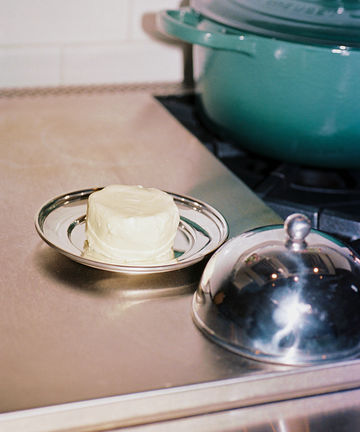 A round pat of butter rests on the Nell Butter Dish with a lid, set on a metal stovetop beside a green pot.