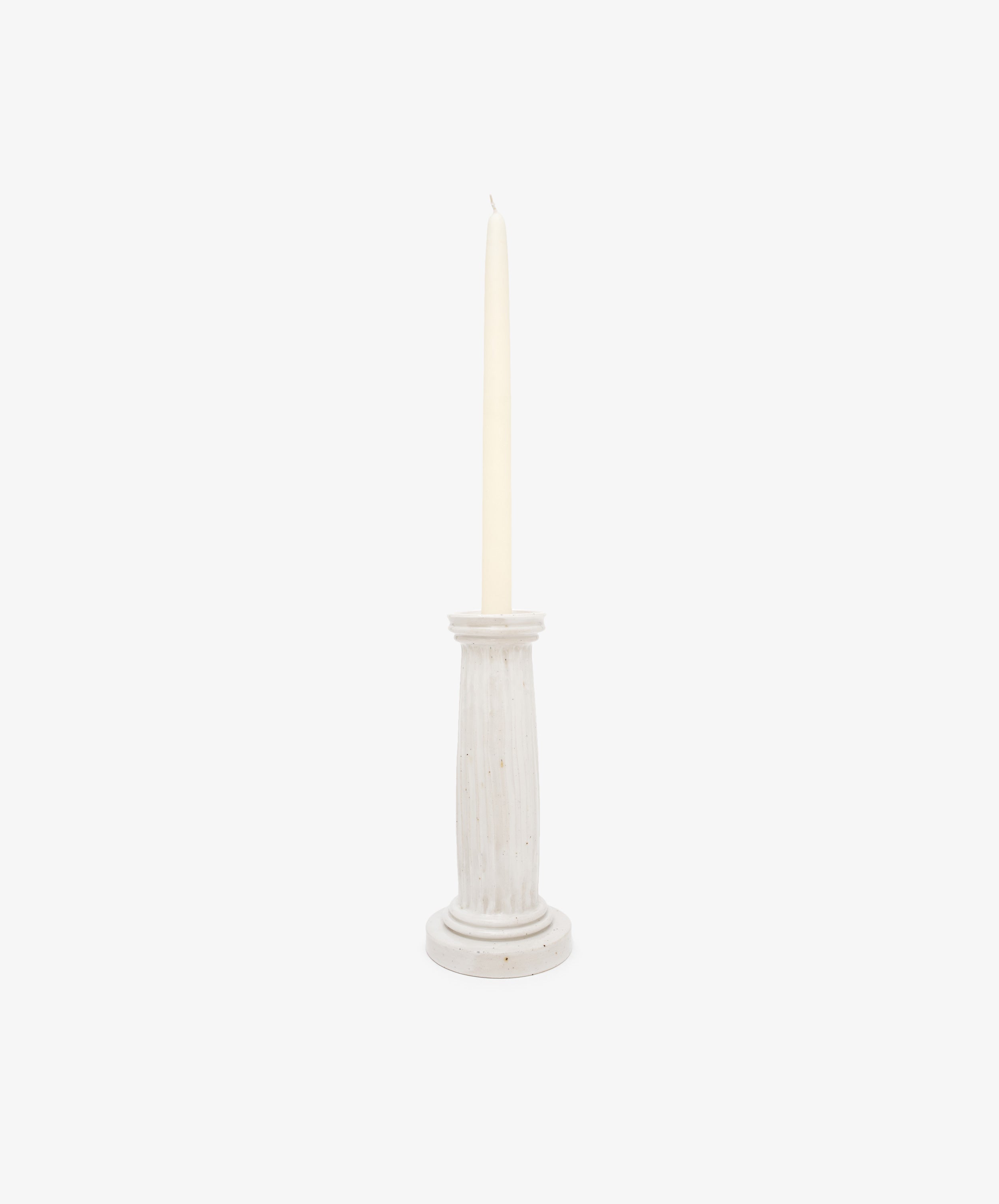 A tall, unlit cream taper candle stands upright in the Column Candleholder, a simple white ceramic piece inspired by sculptural stoneware design, against a plain white background.