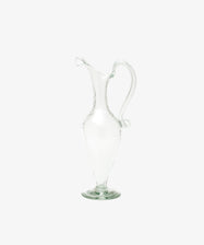 The Alba Vase is a clear glass vessel with a curved handle and narrow spout, standing upright on a round base against a white background—highlighting its artisanal craftsmanship.
