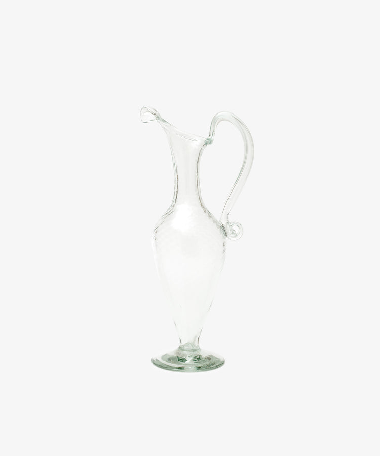 The Alba Vase is a clear glass vessel with a curved handle and narrow spout, standing upright on a round base against a white background—highlighting its artisanal craftsmanship.