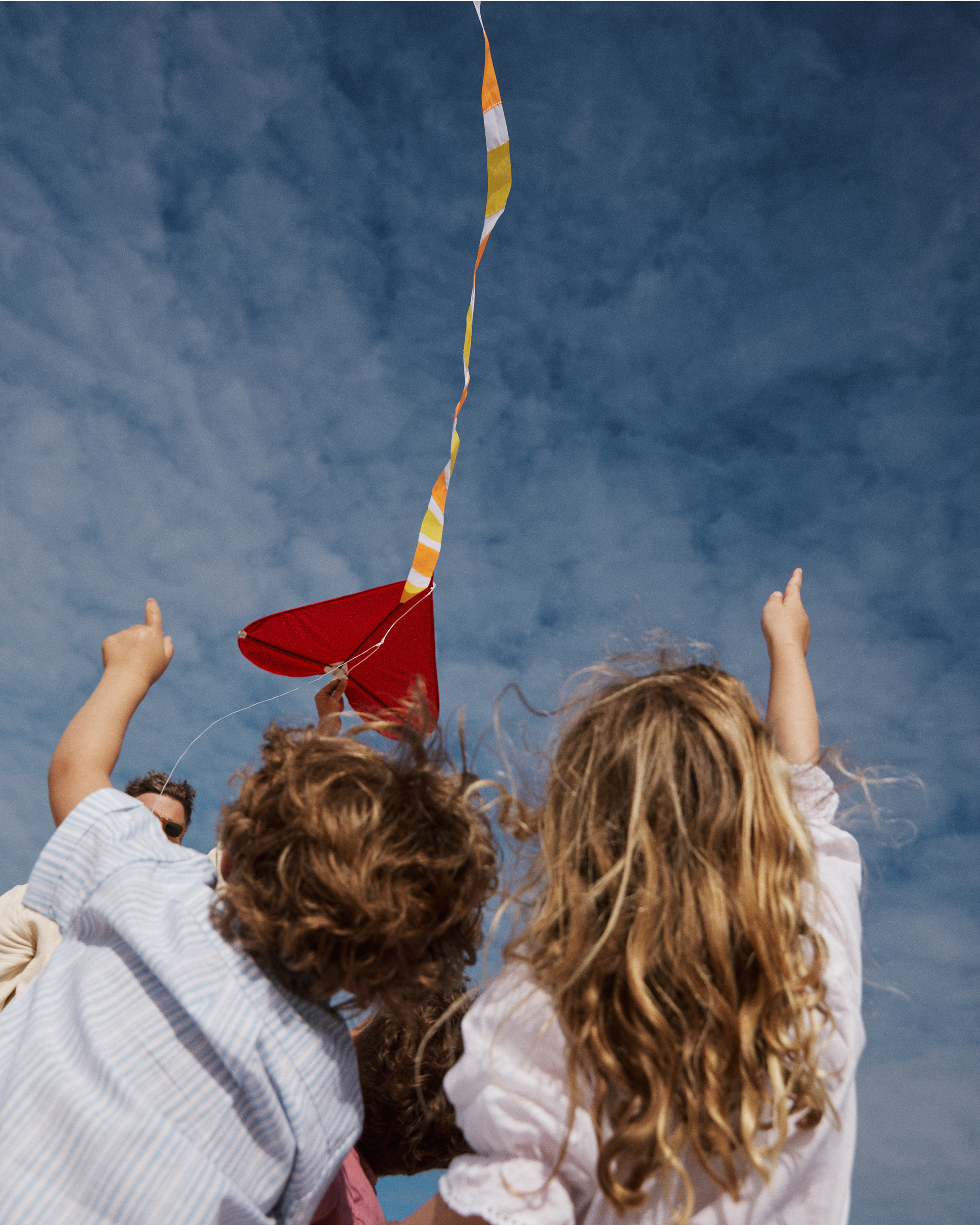 Two children with light brown hair are flying a red Love Supreme Kite under a blue sky with scattered clouds, both pointing upward toward their soaring gift.