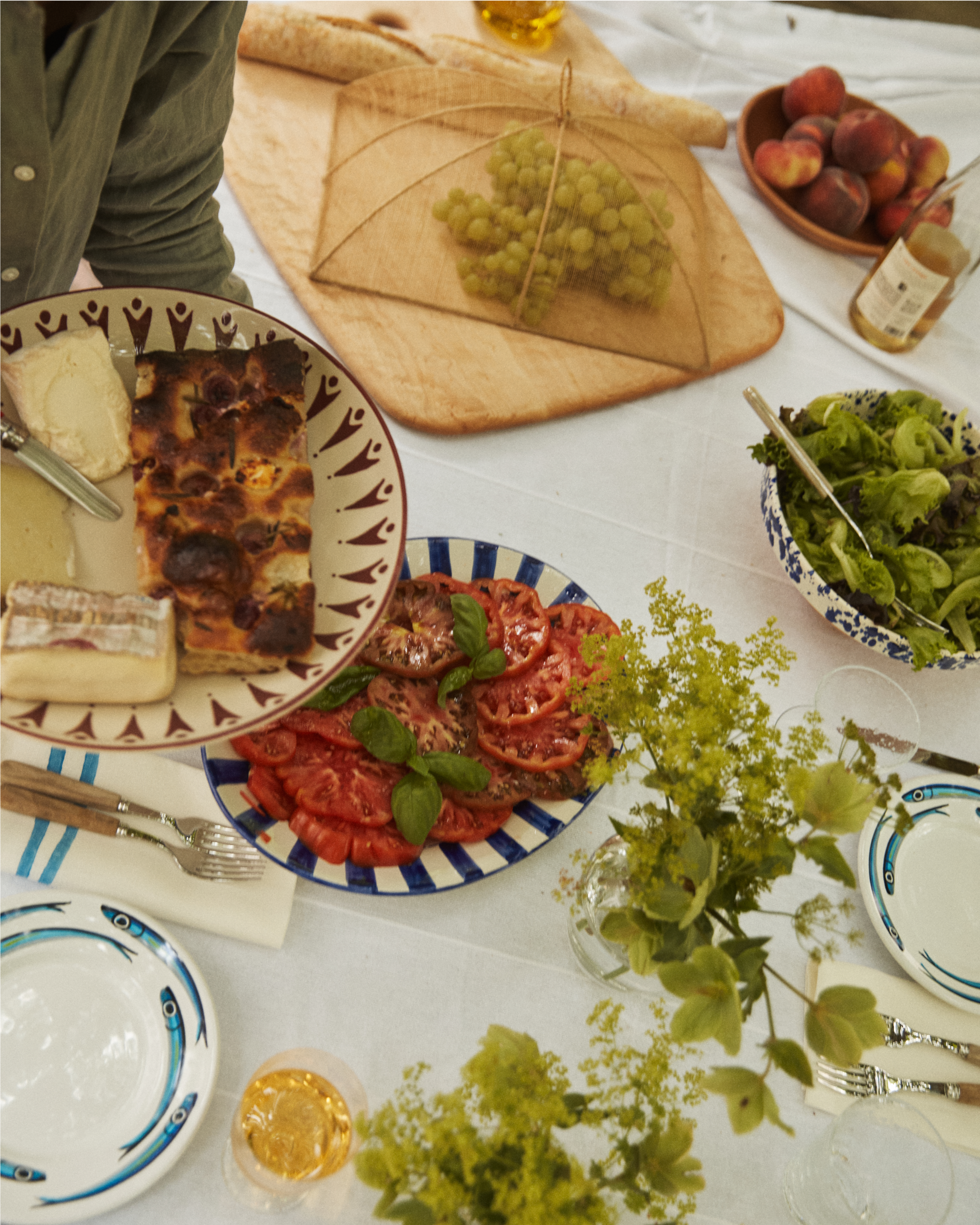 A table set with a Vito Serving Platter holding salad, sliced tomatoes with basil, grapes under a mesh cover, cheese, bread, and drinks. A person uses handmade serveware to serve food onto a plate.