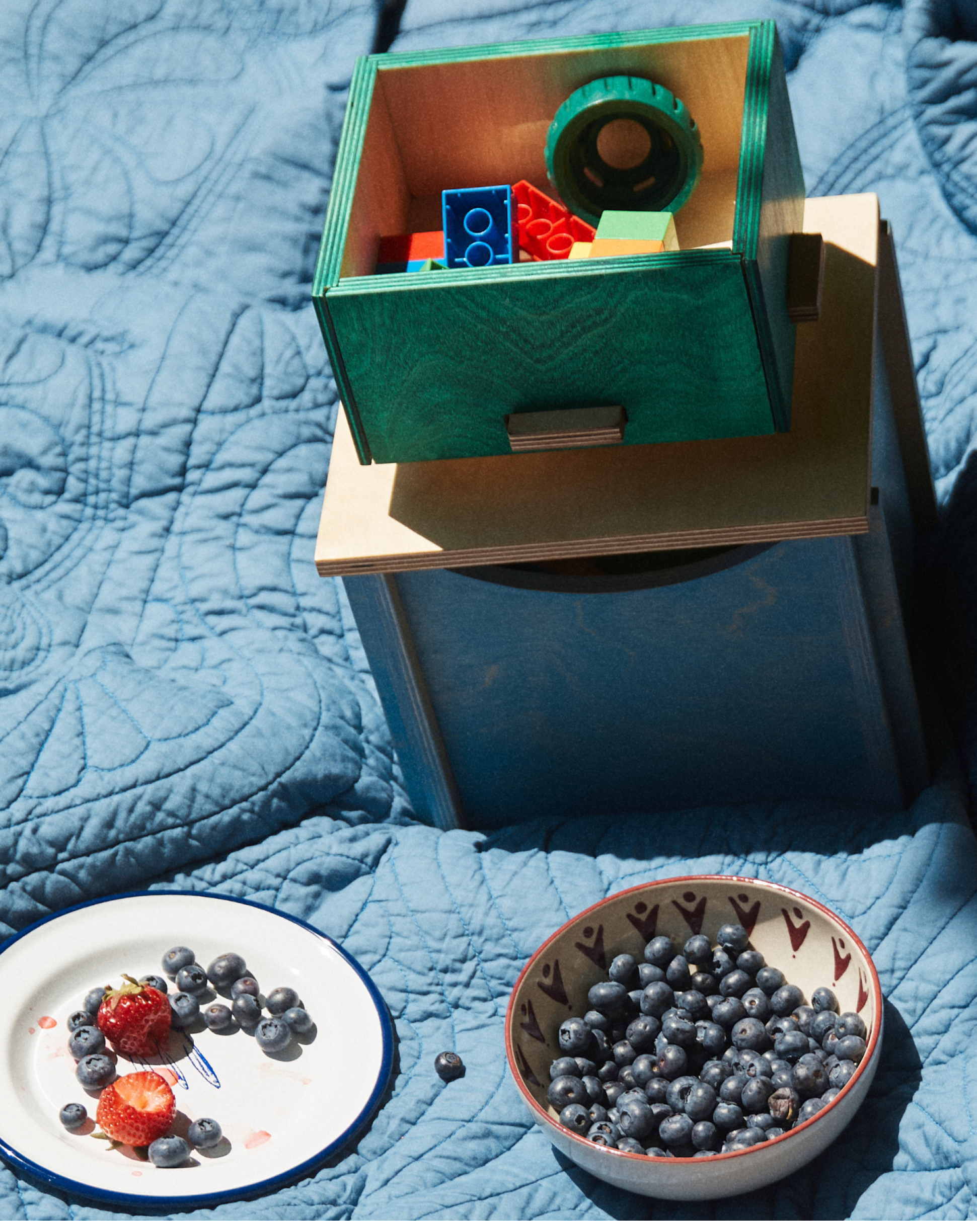 A plate with strawberries and blueberries, a bowl of blueberries, and a Milo Keepsake Box filled with toy blocks sit on a blue quilted blanket.