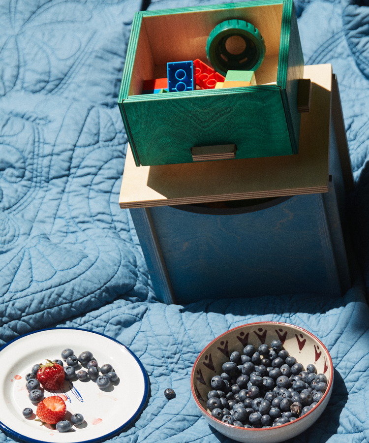 A plate with strawberries and blueberries, a bowl of blueberries, and a Milo Keepsake Box filled with toy blocks sit on a blue quilted blanket.