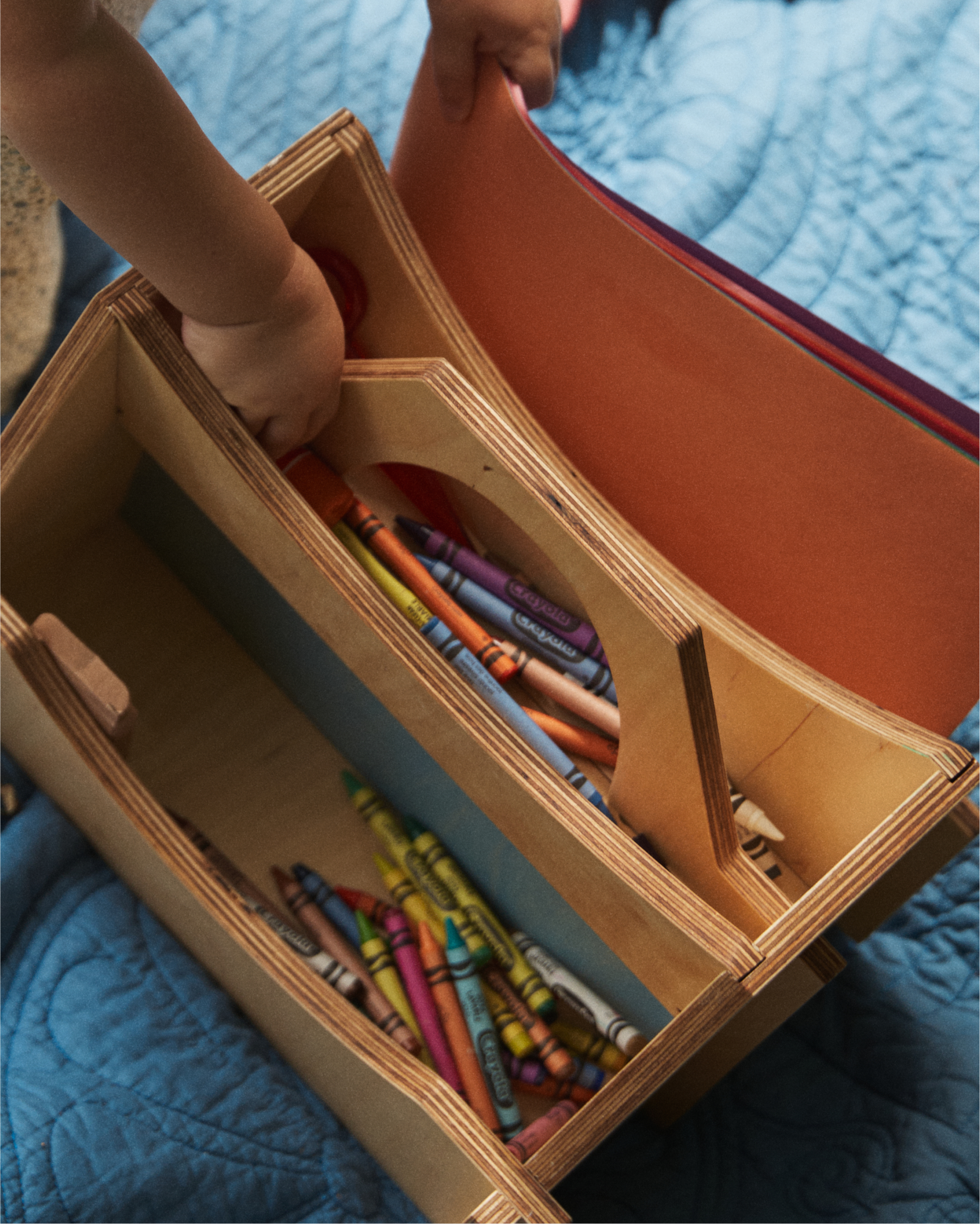 A child places construction paper into the Casper Caddy, a wooden organizer filled with crayons and art supplies, resting on a blue quilt—a perfect kids storage solution.