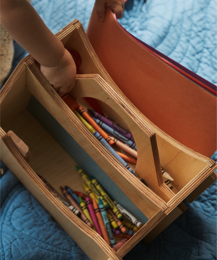 A child places construction paper into the Casper Caddy, a wooden organizer filled with crayons and art supplies, resting on a blue quilt—a perfect kids storage solution.