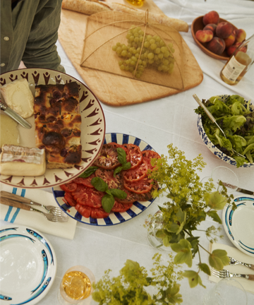 A table set with a Vito Serving Platter holding salad, sliced tomatoes with basil, grapes under a mesh cover, cheese, bread, and drinks. A person uses handmade serveware to serve food onto a plate.