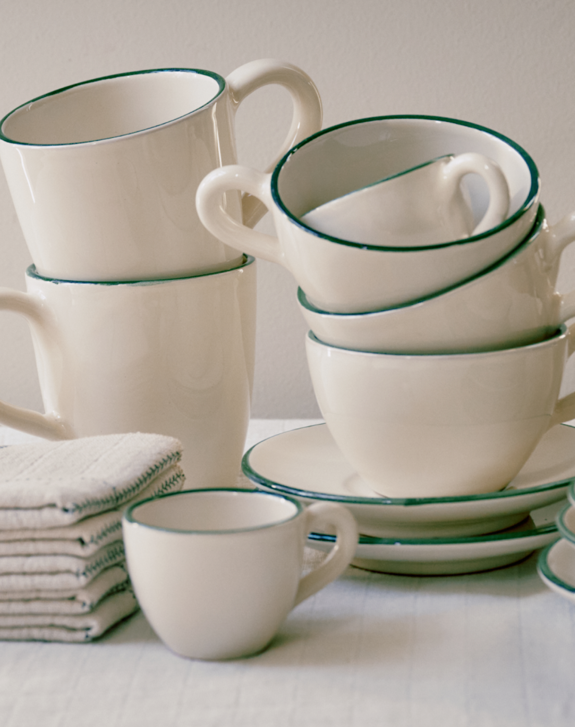 The Guido Coffee Bundle, featuring white ceramic coffee cups and saucers with green rims plus matching plates, is displayed beside a neatly folded stack of beige napkins on a light surface.