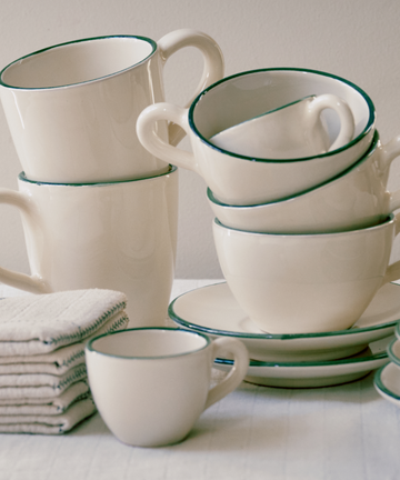 The Guido Coffee Bundle, featuring white ceramic coffee cups and saucers with green rims plus matching plates, is displayed beside a neatly folded stack of beige napkins on a light surface.