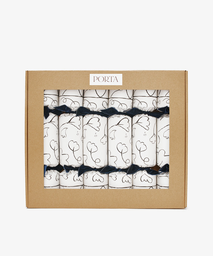 A box of six Capriccio Christmas Crackers featuring white wraps with black line art and navy ribbons, packaged in a labeled brown cardboard window box—ideal festive favors for your holiday party.