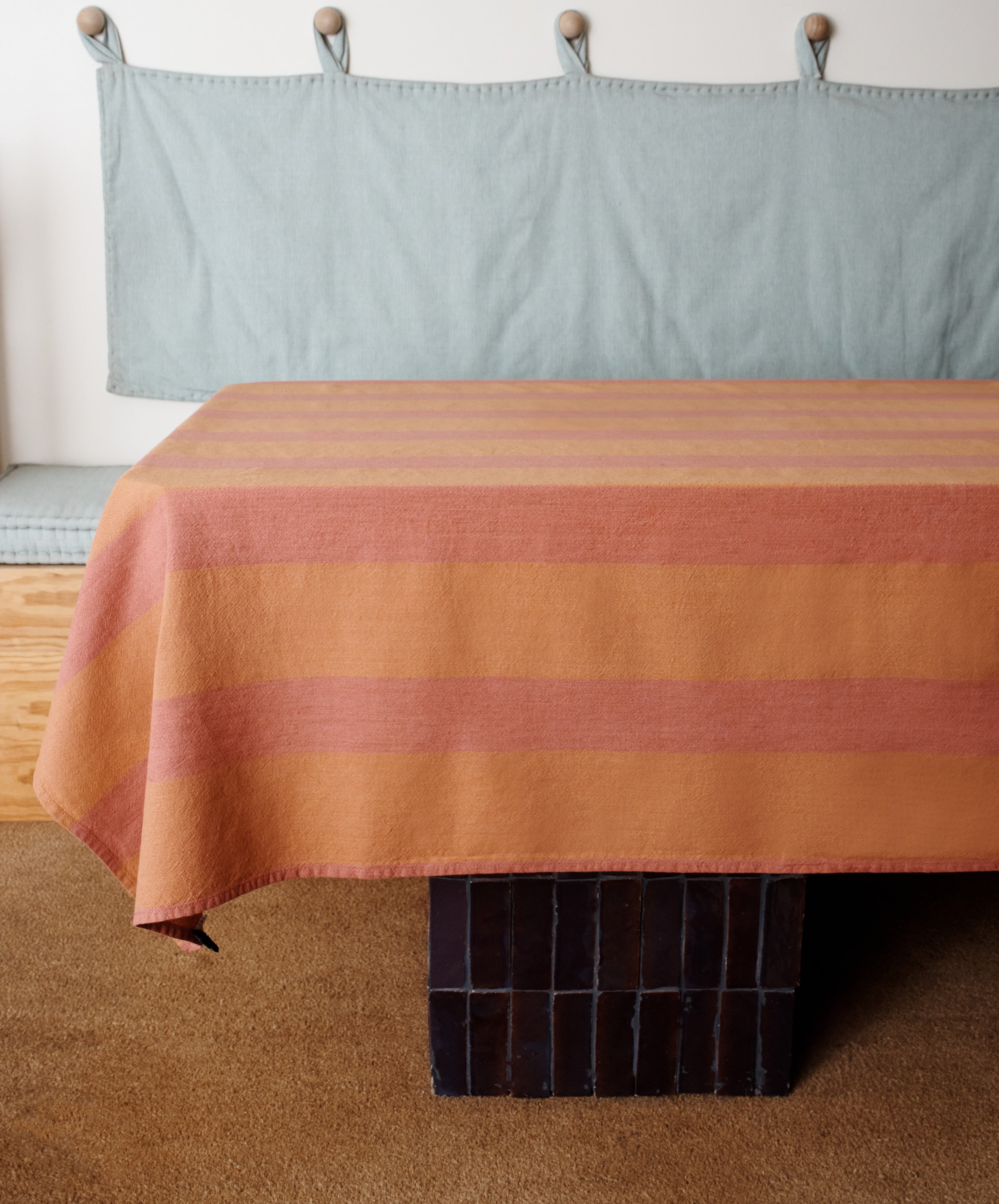 A table with a Cruz Tablecloth in terracotta linen is set on a brown tiled base, with a cushioned bench and hooks in the background.