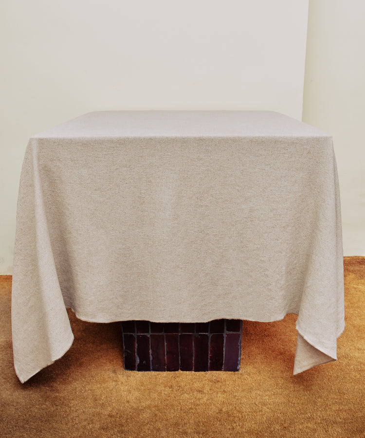 A square table with a Corte Tablecloth in plain beige stands on a brown carpeted floor against a cream-colored wall.
