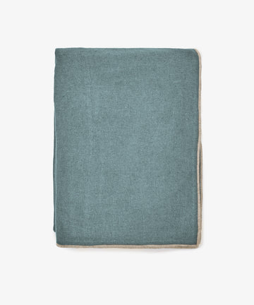The Corte Tablecloth, a neatly folded rectangular piece in teal with beige trim, exudes sophistication as it is displayed against a plain white background.