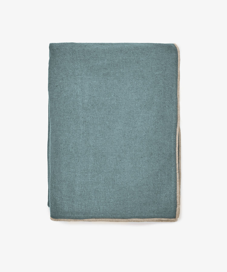 The Corte Tablecloth, a neatly folded rectangular piece in teal with beige trim, exudes sophistication as it is displayed against a plain white background.