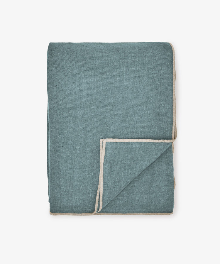 A folded blue Corte Tablecloth with beige trim, displayed on a plain white background, adds a touch of sophistication to any space.