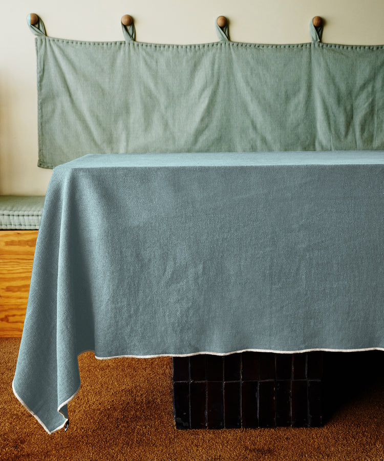 A Corte Tablecloth in light blue covers a table on a brown carpet, complemented by a cushioned bench and hooks for added sophistication.