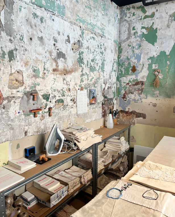 Workshop with weathered walls, shelves of folded fabrics, and an iron on a workbench