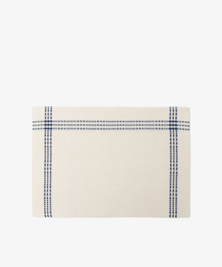 The Tilde Placemat, Set of 6, features a rectangular cream cotton-linen design with two intersecting blue stripes forming a cross near the edges, inspired by the classic Daldräll pattern and displayed on a white background.