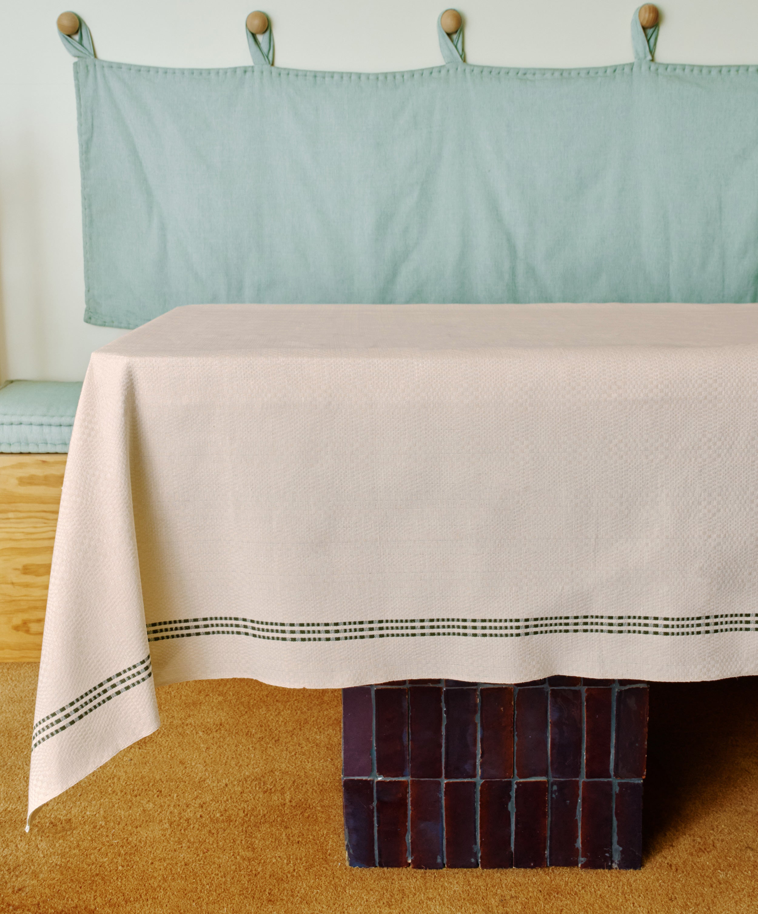A table with the Tilde Tablecloth, featuring cream and black Swedish cotton-linen stripes, stands on a brown brick base, with a light blue cushioned bench and hooks visible in the background.