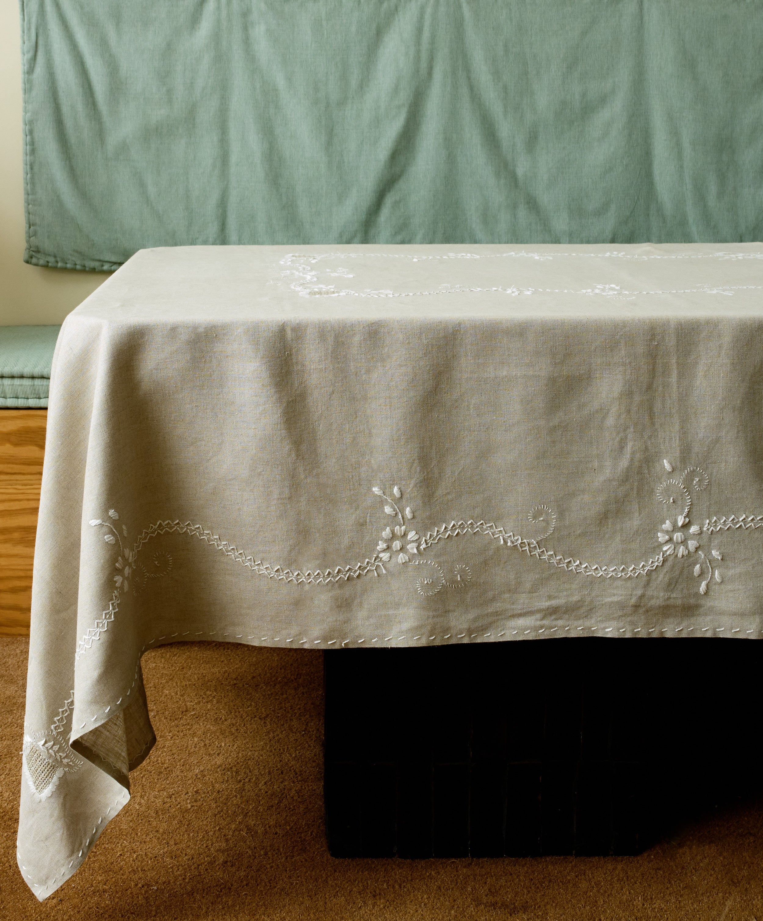 Zelia Tablecloth in natural linen with white embroidery details, draped over table in studio.