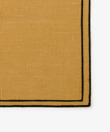 A close-up view of the Flavia Napkin set of 6 in tan woven fabric, featuring two thin black border lines and a detailed double-line stitch, highlighting the texture and corner of the napkin.
