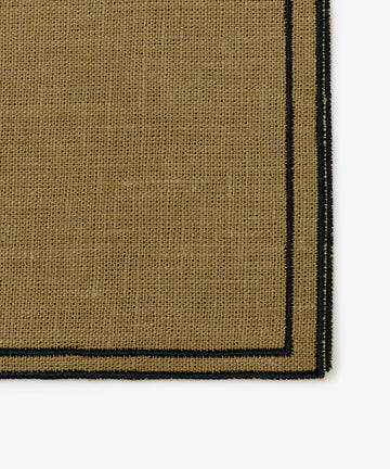 Close-up of the corner of a Flavia Napkin, Set of 6—rectangular tan woven fabric with a black border and double-line stitch, evoking fine linen napkins.