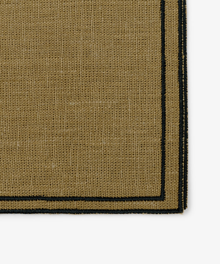 Close-up of the corner of a Flavia Napkin, Set of 6—rectangular tan woven fabric with a black border and double-line stitch, evoking fine linen napkins.