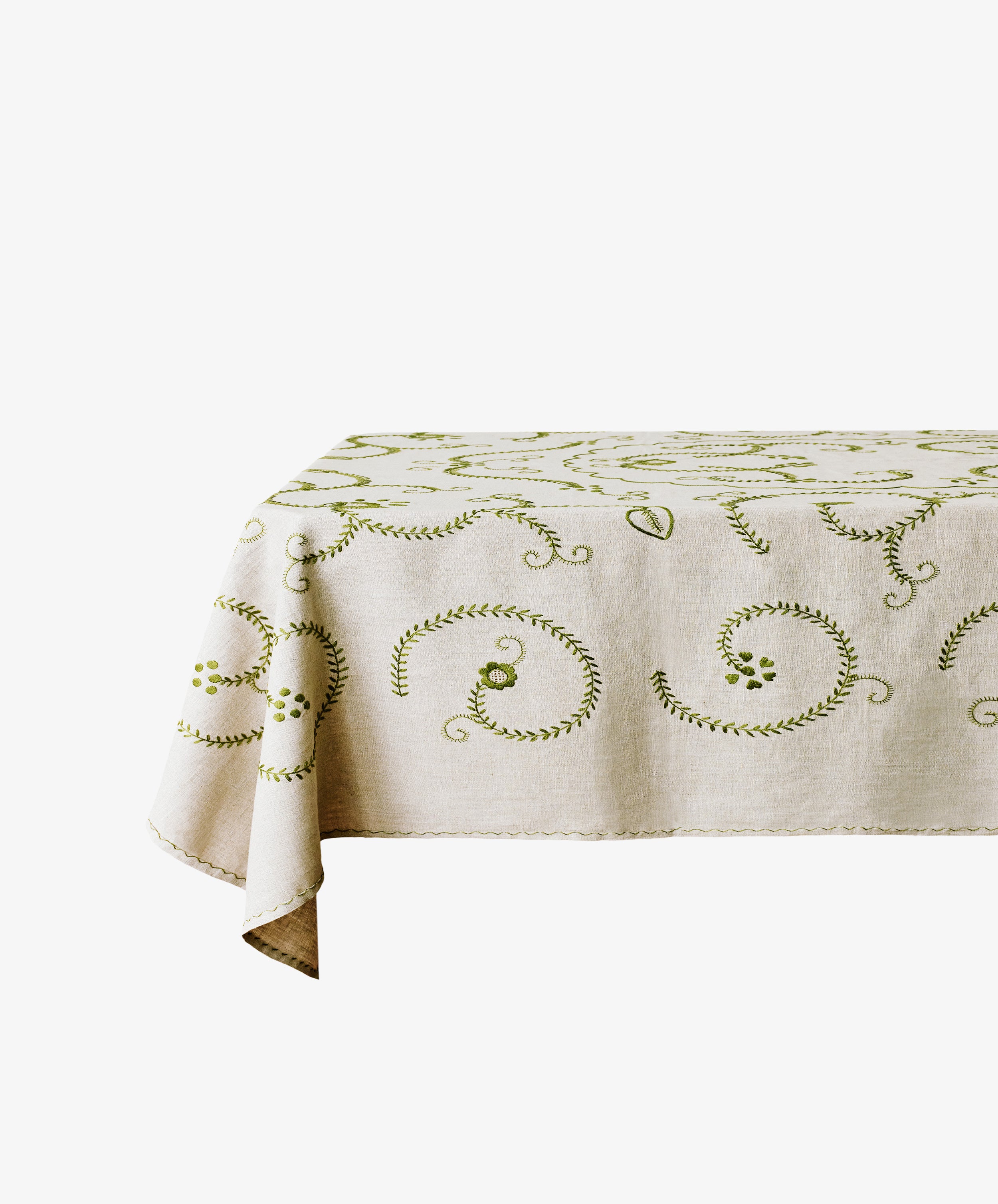 The Inez Tablecloth, made of raw linen with green hand-embroidered swirl and leaf patterns, is draped over a rectangular table against a white background.