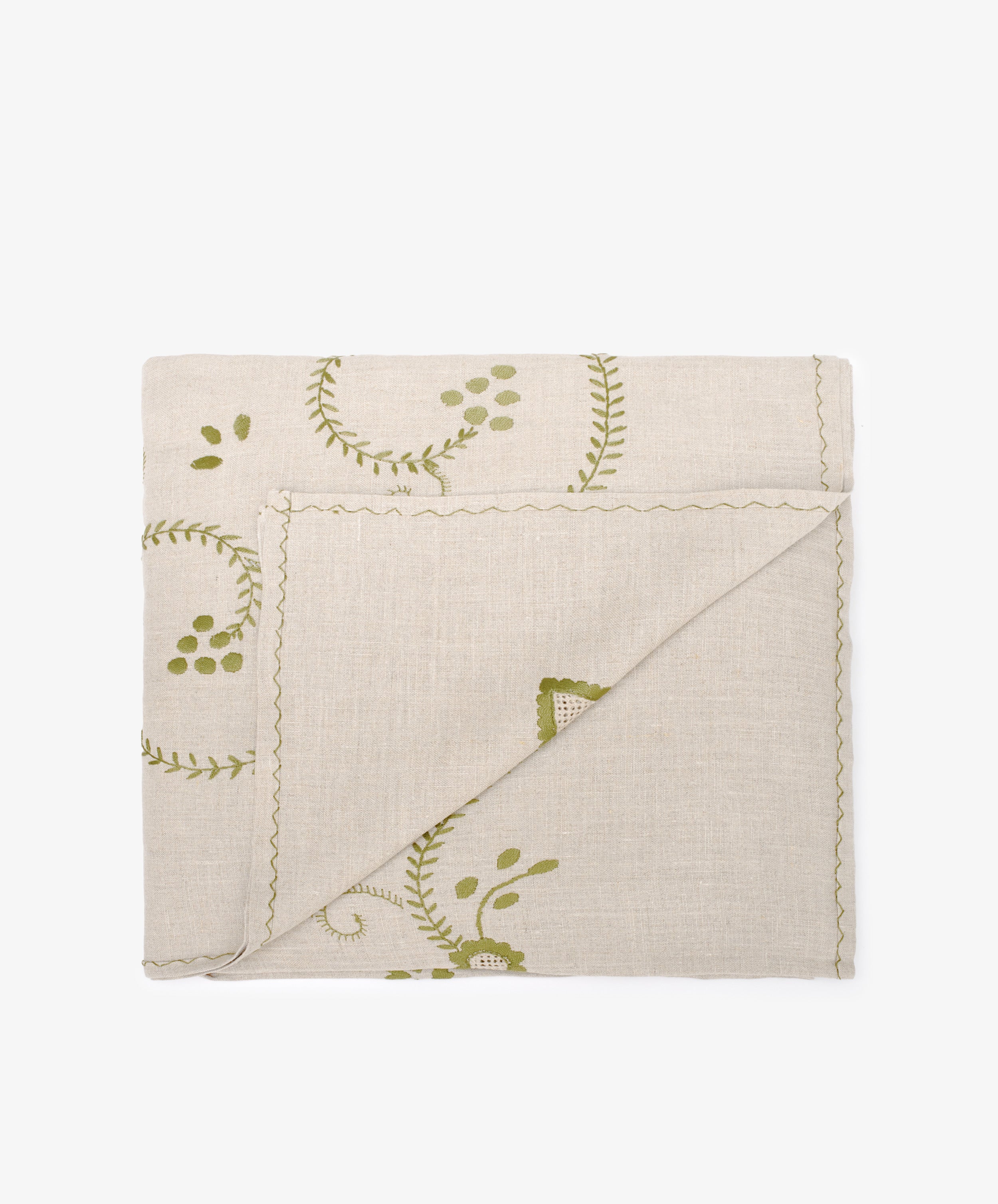 The Inez Tablecloth features a beige fabric with green leaf and vine patterns, scalloped edges, and hand-embroidered-style detailing, shown folded on a plain white background.