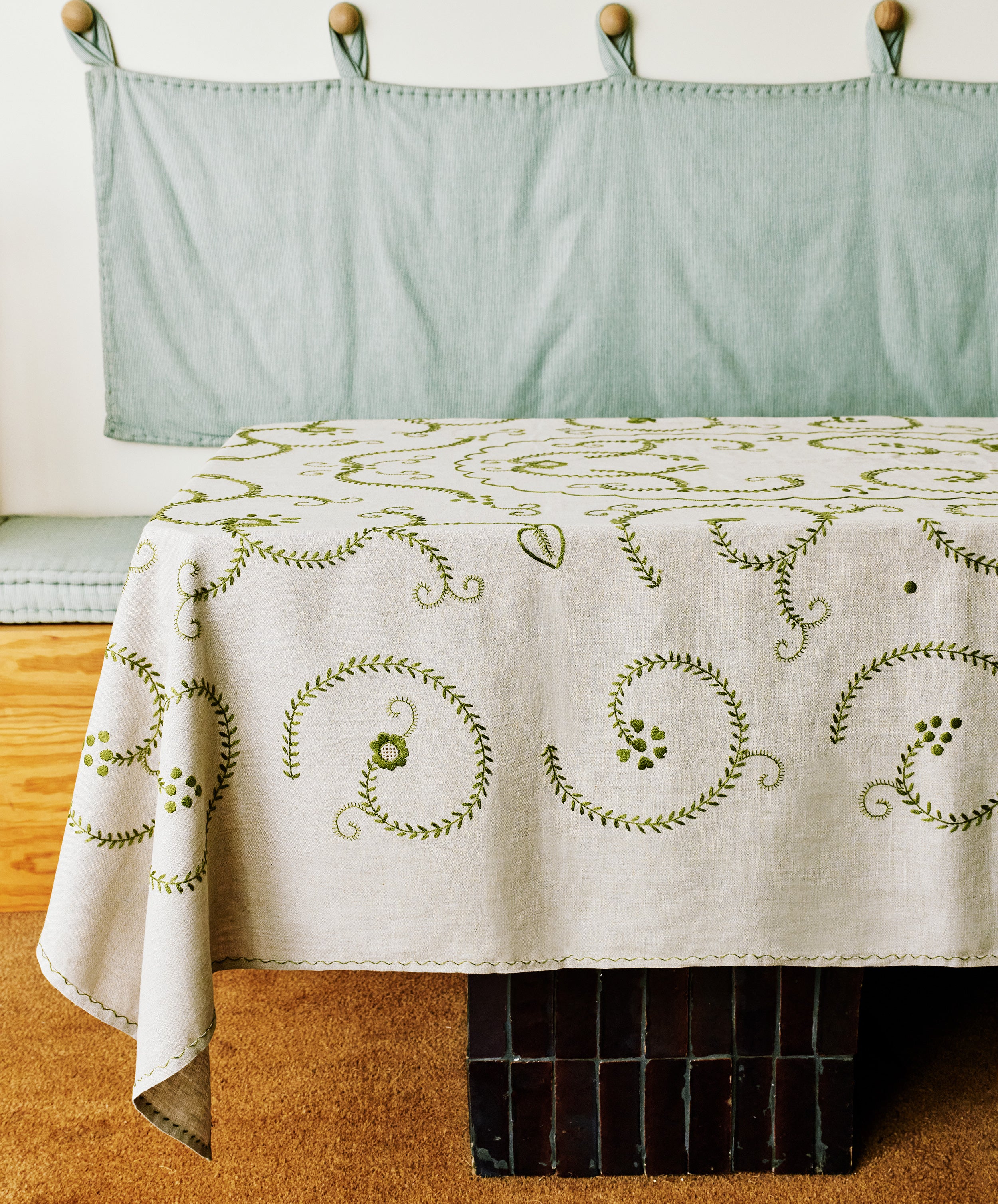 A rectangular table covered with the Inez Tablecloth, a white Portuguese linen featuring green embroidered swirls, stands before a cushioned bench with a light blue fabric backrest.