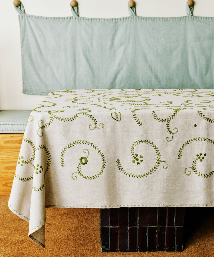 A rectangular table covered with the Inez Tablecloth, a white Portuguese linen featuring green embroidered swirls, stands before a cushioned bench with a light blue fabric backrest.