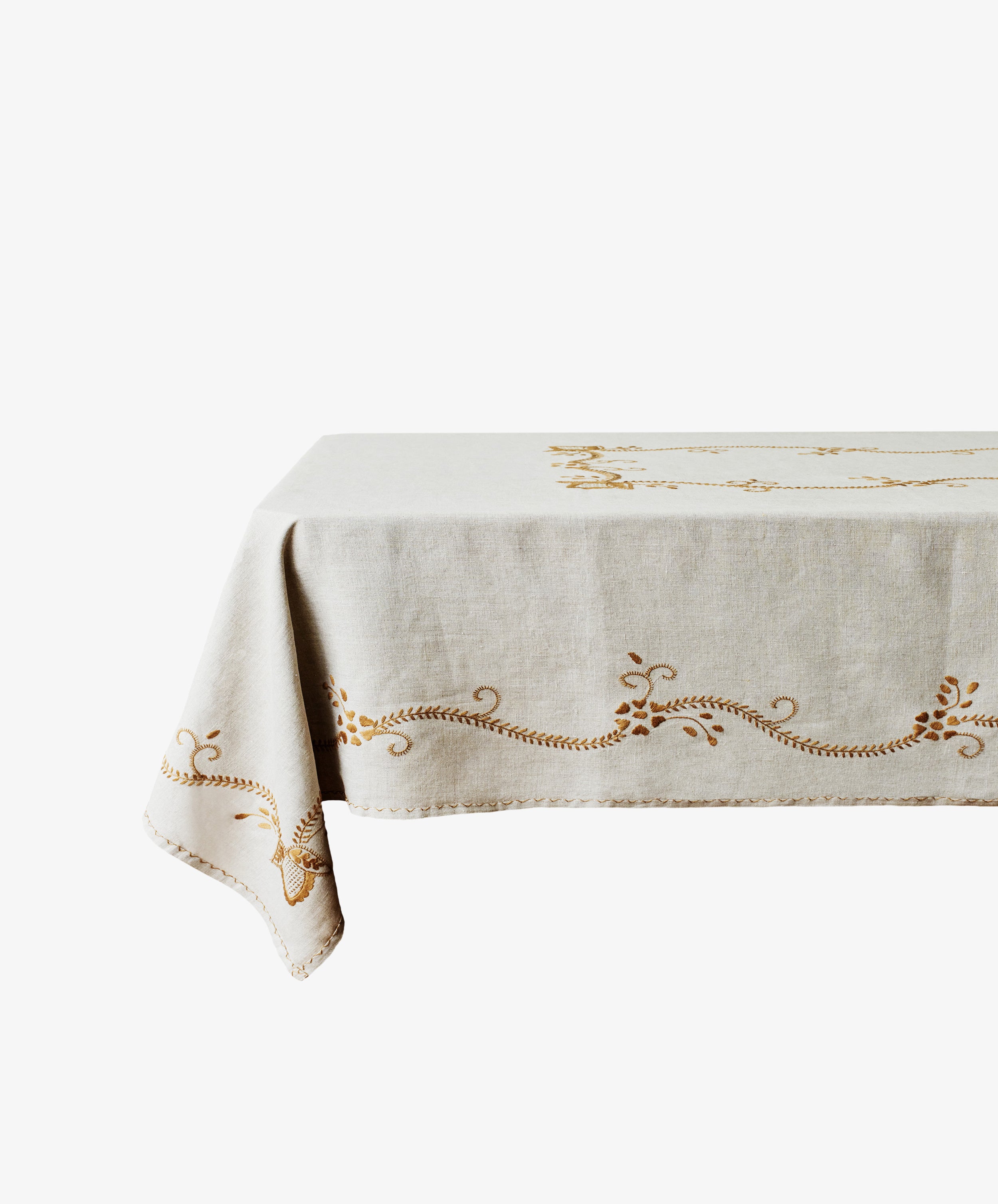 The Zelia Tablecloth, a beige linen piece with embroidered gold patterns, is draped over a rectangular table set against a white background.