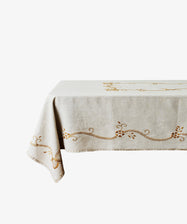 The Zelia Tablecloth, a beige linen piece with embroidered gold patterns, is draped over a rectangular table set against a white background.