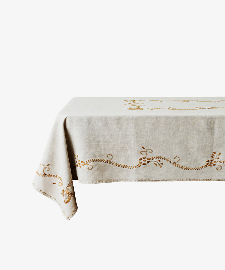 The Zelia Tablecloth, a beige linen piece with embroidered gold patterns, is draped over a rectangular table set against a white background.