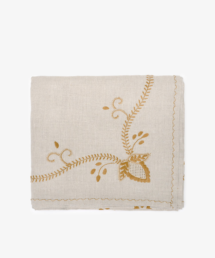 The Zelia Tablecloth is a folded beige linen piece featuring mustard yellow hand-embroidered floral and leaf patterns along one corner and edge.