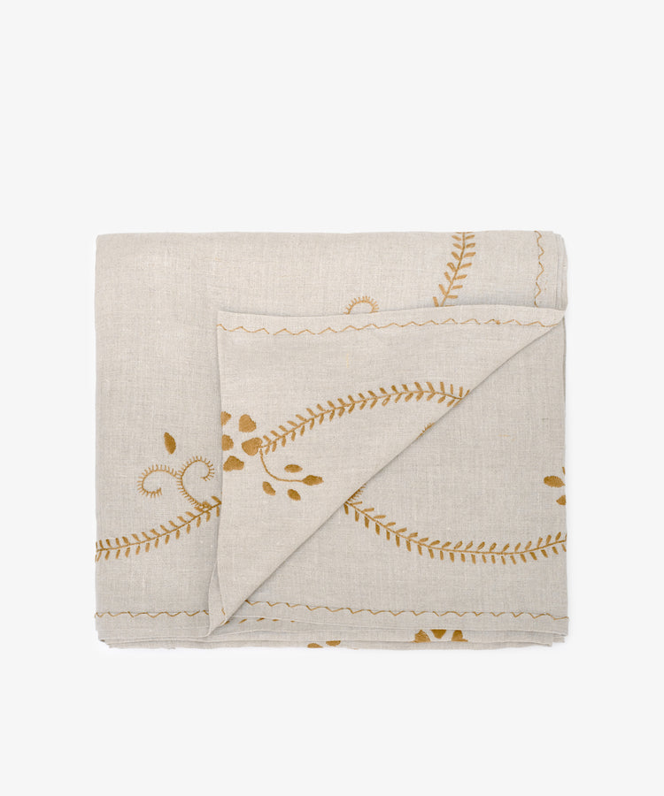 The Zelia Tablecloth is a folded beige linen piece with yellow embroidered leaf and vine patterns, echoing traditional Portuguese designs, displayed on a white background.