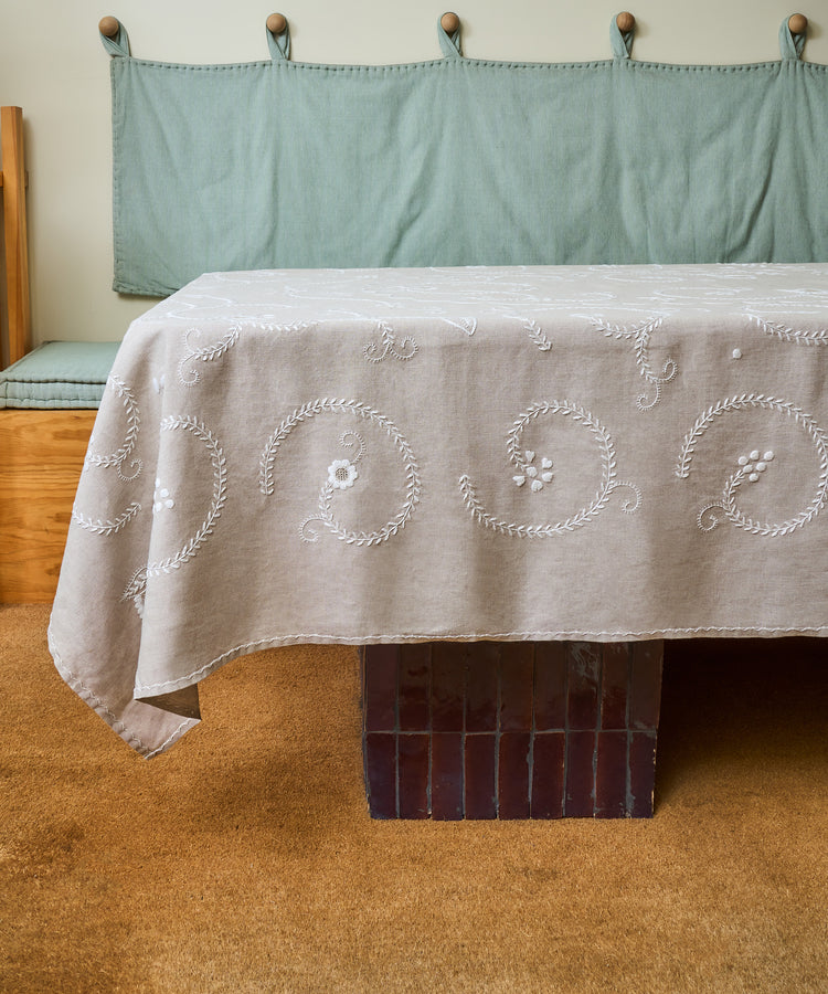 A table draped with the Inez Tablecloth stands on a dark brown tiled base beside a bench with a light green cushion and backrest, set atop a tan carpet.