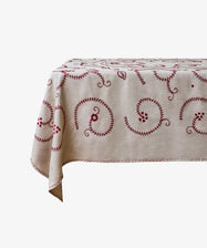 The Inez Tablecloth, made from Portuguese raw linen with red hand-embroidered swirling patterns, is draped over a rectangular table set against a white background.